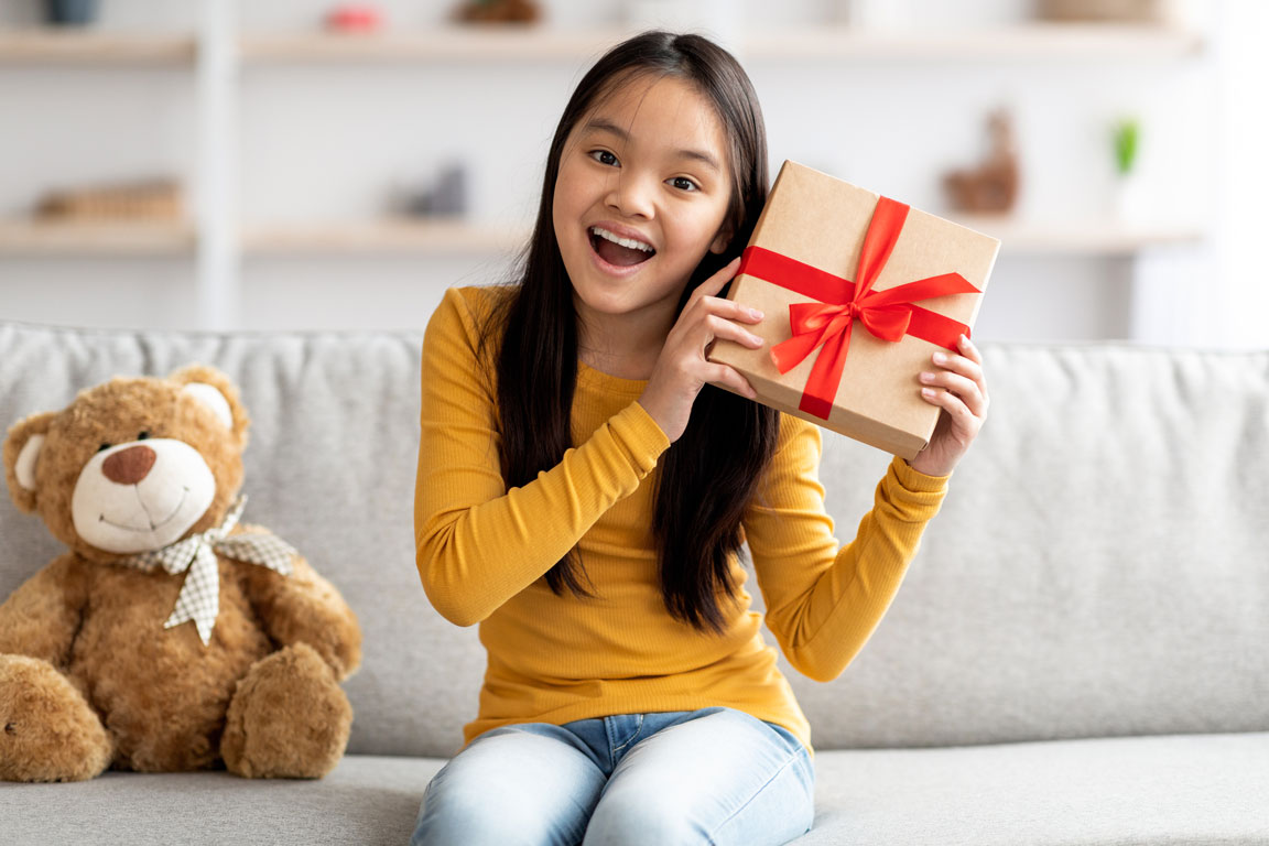 young girl holding holiday gift while sitting on couch next to a teddy bear